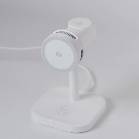 A demonstration video of the white Schlüssel 3-in-1 wireless charging station. A hand sequentially places an Apple Watch on the top dock, AirPods on the base, and an iPhone horizontally on the front magnetic Qi2 pad, showing all three devices charging simultaneously