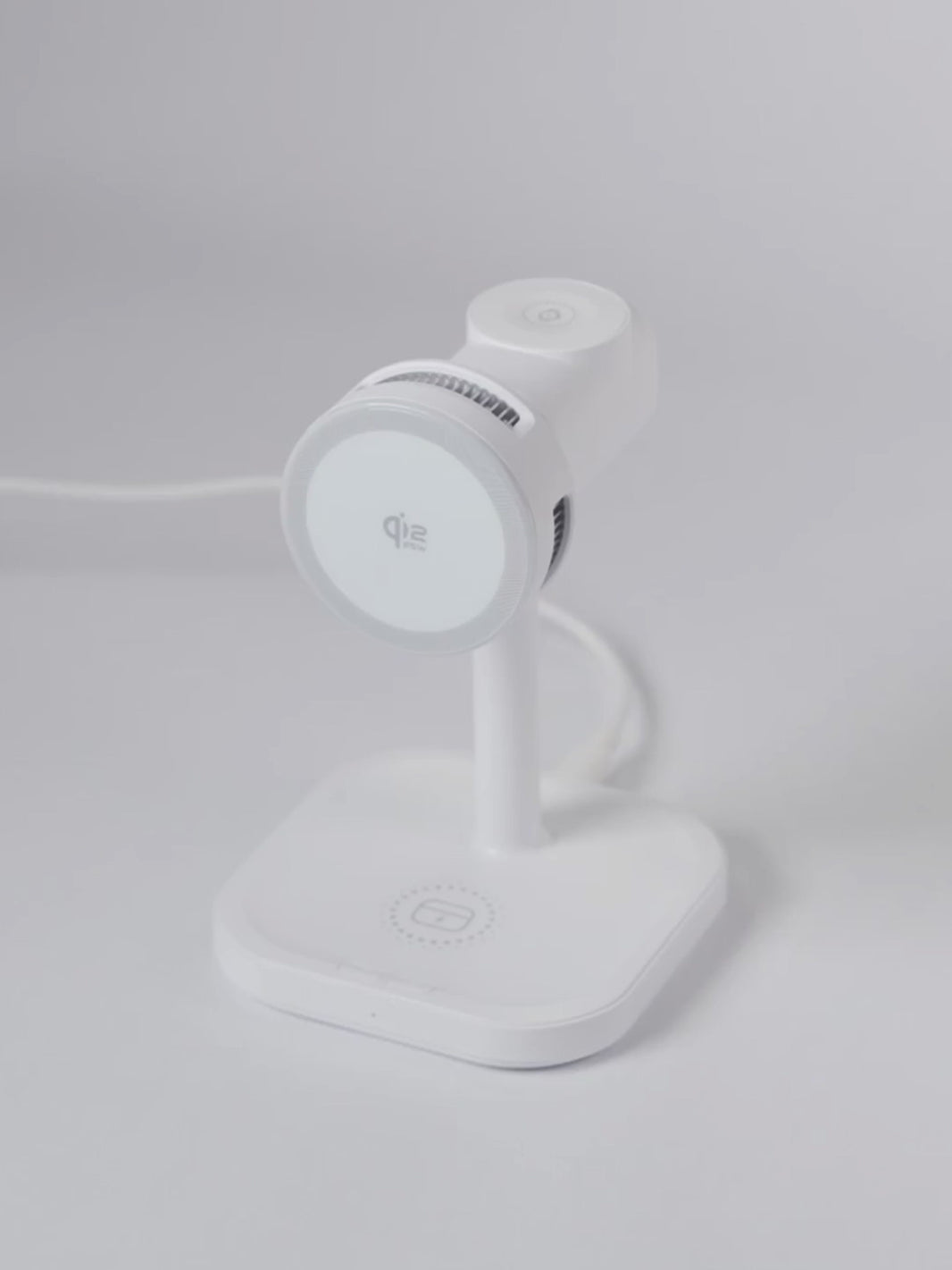 A demonstration video of the white Schlüssel 3-in-1 wireless charging station. A hand sequentially places an Apple Watch on the top dock, AirPods on the base, and an iPhone horizontally on the front magnetic Qi2 pad, showing all three devices charging simultaneously
