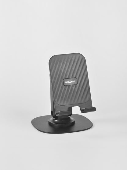A standalone black Schlüssel adjustable desktop phone stand featuring a vertically ridged backrest and a wide, flat base plate, captured from a three-quarter angle against a neutral gray background