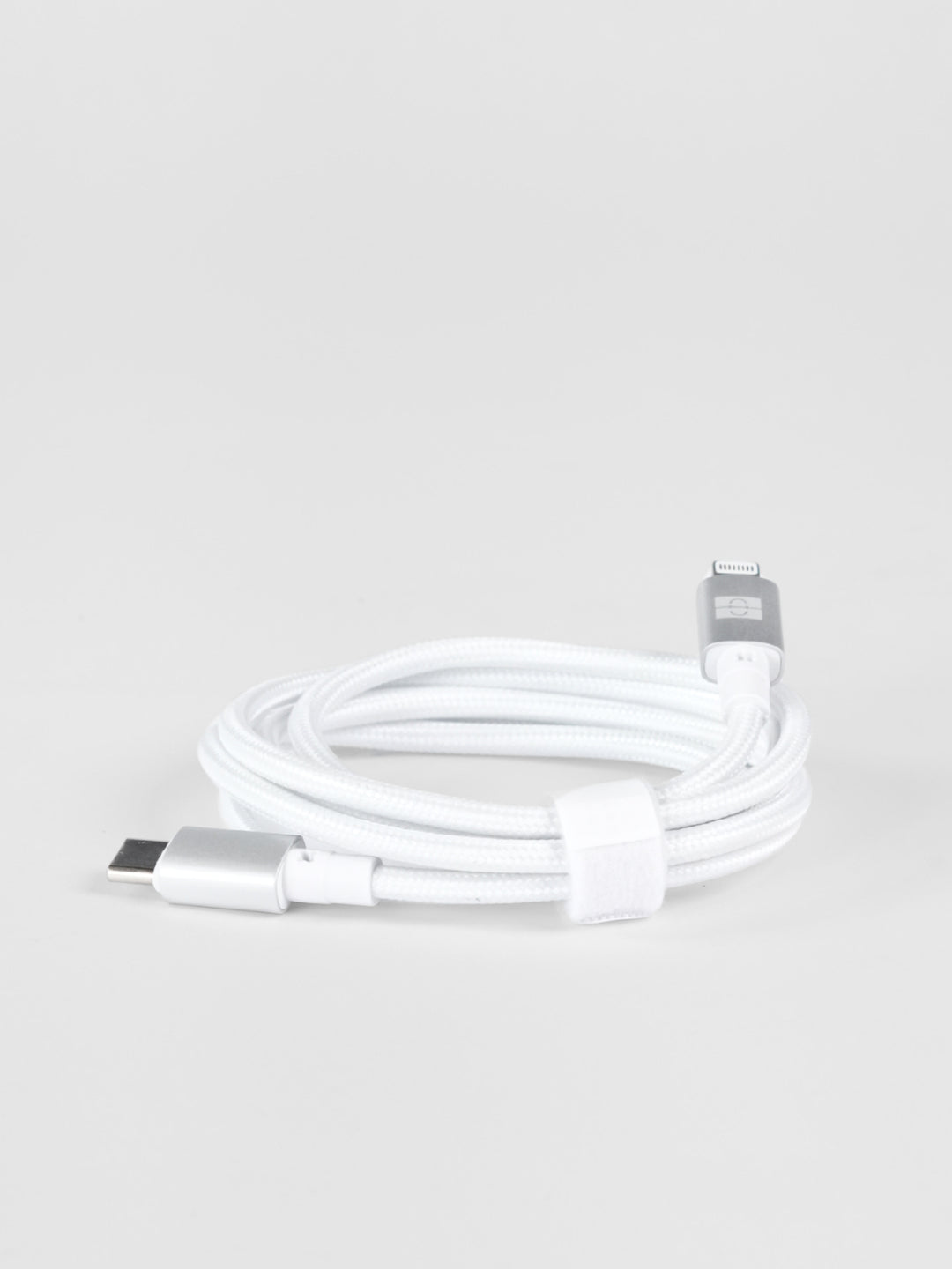 A neatly coiled white braided Schlüssel USB-C to Lightning cable featuring premium silver aluminum connector housings, fastened with a white hook-and-loop strap on a light gray background